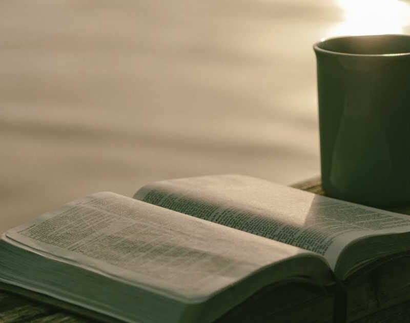 Cup of coffee and bible on dock rail of lake with sunrise and water in the background.
