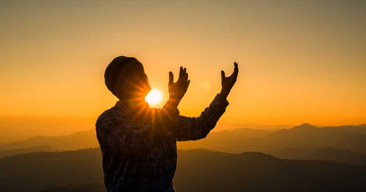 Man standing with praying hands outstretched at sunrise.