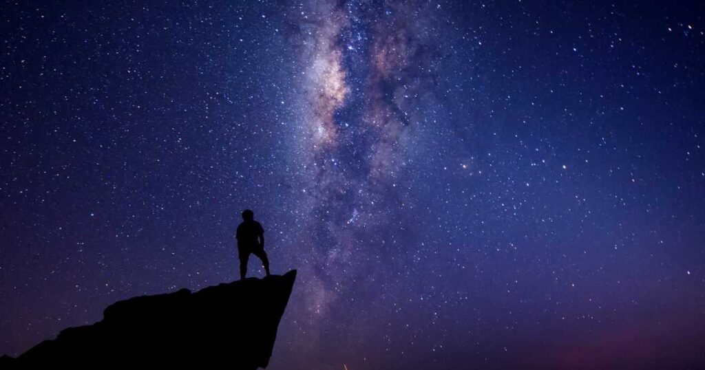 Man standing on edge of cliff at night with Milky Way Galaxy in the background.