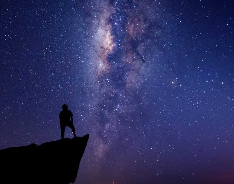 Man standing on edge of cliff at night with Milky Way Galaxy in the background.